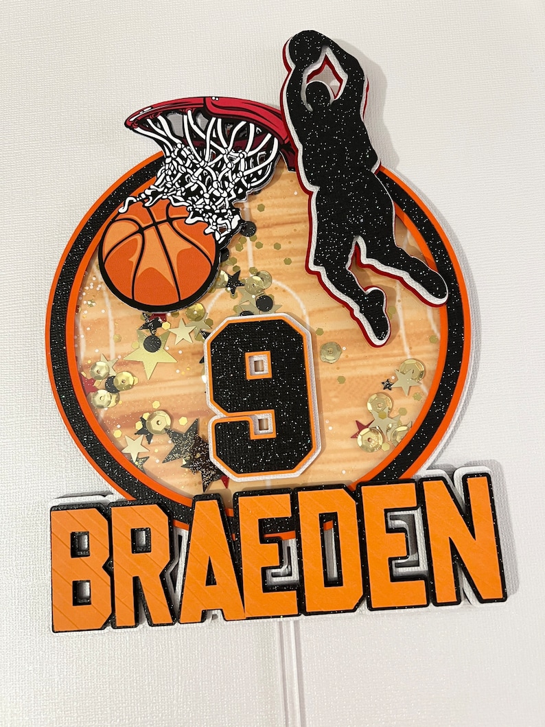 May include: Orange and black basketball themed cake topper with a basketball going through a hoop, a silhouette of a player dunking, the number 9, and the name "Braeden".