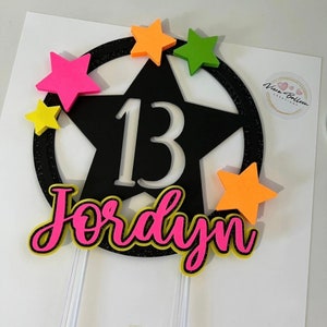 May include: A black star-shaped cake topper with the number 13 in white. The name "Jordyn" is written in pink with a yellow outline. The topper is decorated with colorful stars in pink, yellow, green, and orange.