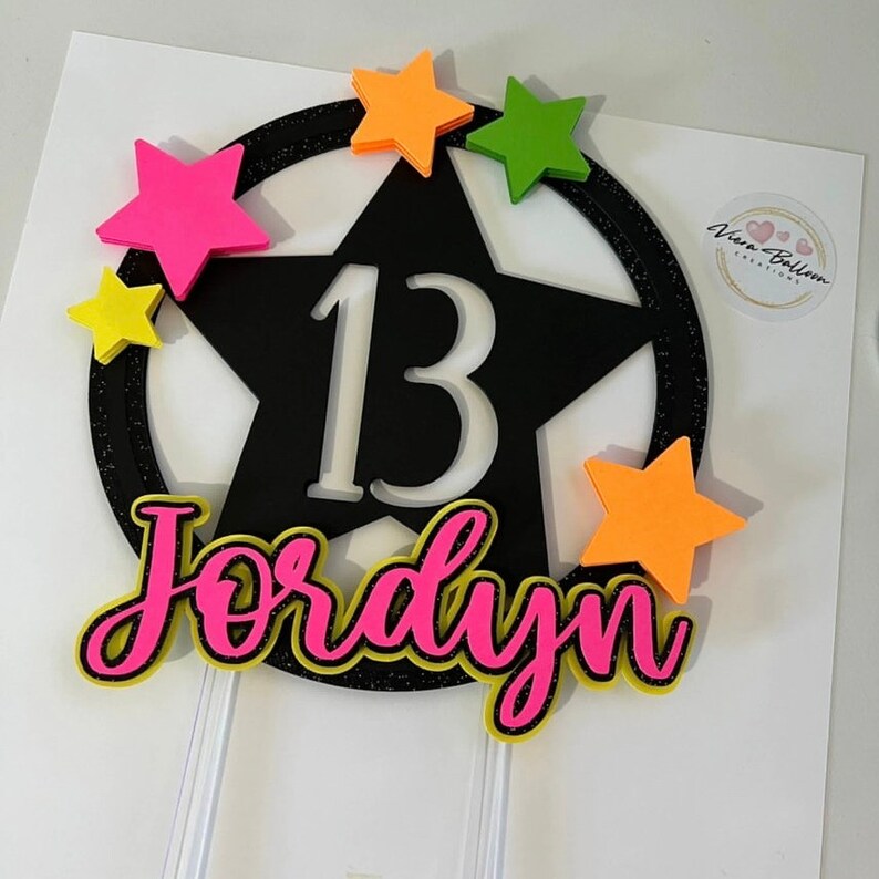 May include: A black star-shaped cake topper with the number 13 in white. The name "Jordyn" is written in pink with a yellow outline. The topper is decorated with colorful stars in pink, yellow, green, and orange.