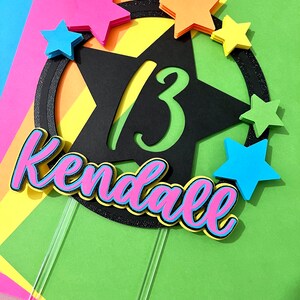 May include: A colorful cake topper with the name "Kendall" in pink and blue, and the number "13" in green. The topper is decorated with colorful star shapes and a black star outline.