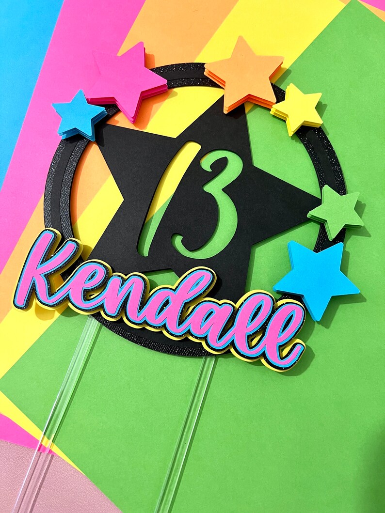 May include: A colorful cake topper with the name "Kendall" in pink and blue, and the number "13" in green. The topper is decorated with colorful star shapes and a black star outline.
