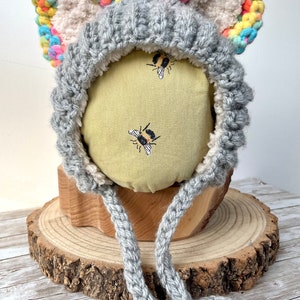 May include: A grey crocheted bonnet featuring two large animal-style ears with multicoloured yarn edges and a cream-coloured fuzzy interior lining. The bonnet includes two long grey crocheted ties hanging from the front and a textured knit pattern.