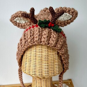 May include: A light brown crocheted hat featuring two dark brown antlers, two large ears with cream-coloured centres, and a decorative band of red yarn accented with green crocheted holly leaves and three small red berries on the front.