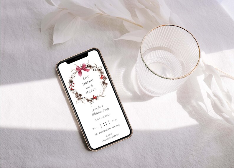 May include: A smartphone displaying a digital invitation for a Christmas party. The invitation features a wreath with red berries and a bow, and the text "Eat Drink and be Happy". The invitation also includes the date, time, and location of the party.