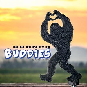 May include: A black textured silhouette of a Bigfoot figure making a heart shape with its hands above its head. The text BRONCO BUDDIES is printed in blue and white, with the text by ThirdDimensionGifts appearing in smaller black font below.