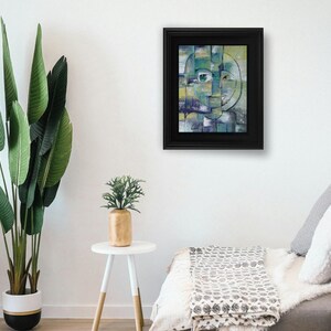 May include: Abstract painting of a face with green, blue, and yellow colors. The painting is framed in a black frame and is hanging on a white wall.