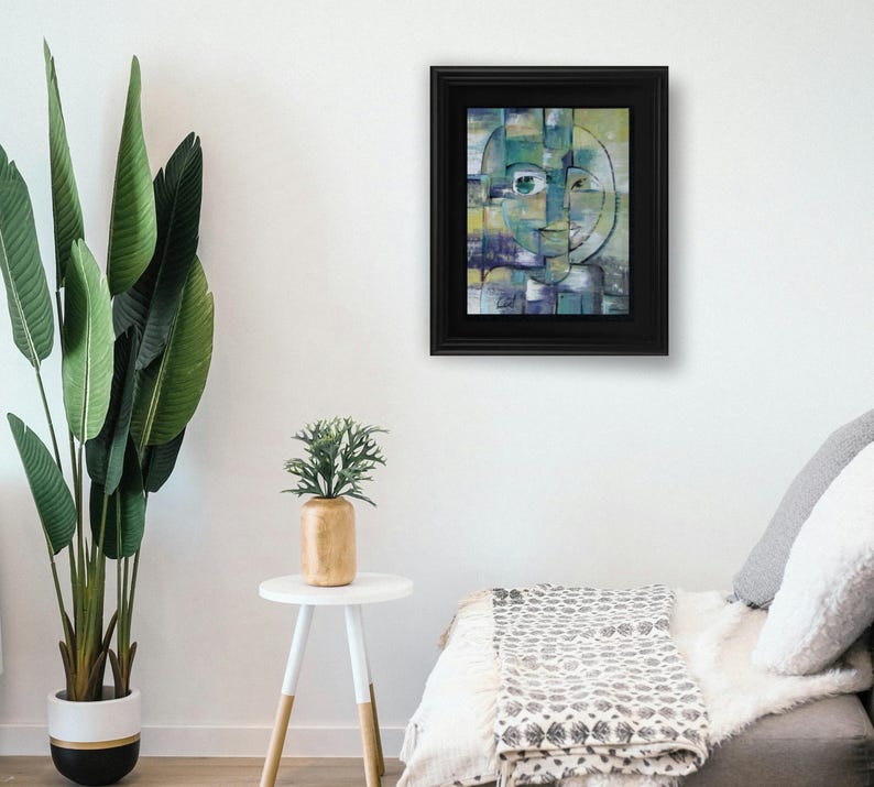 May include: Abstract painting of a face with green, blue, and yellow colors. The painting is framed in a black frame and is hanging on a white wall.