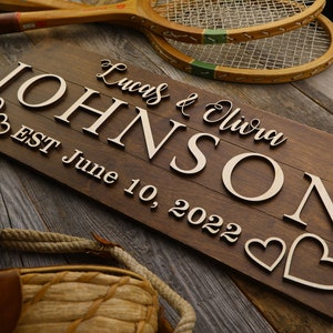 May include: A brown wooden sign with the names "Lucas & Olivia Johnson" and the date "June 10, 2022". The sign has light-coloured lettering and heart shapes. Vintage tennis rackets are in the background.