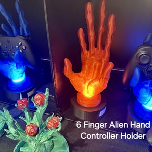 May include: Three 3D printed alien hand controller holders. The hands are blue, orange, and red. The hands are holding video game controllers. The text "6 Finger Alien Hand Controller Holder" is at the bottom of the image.