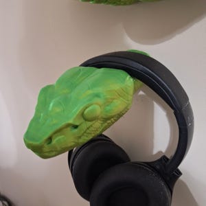 May include: A green snake head-shaped headphone holder with a black headphone set. The snake head is mounted on a wall. The headphones are black with large, round earcups.