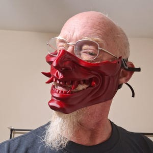 May include: A vibrant red Oni mask with sharp teeth and horns. The mask has a detailed, textured surface and is worn with black straps. The mask is a traditional Japanese demon mask, often used in theater and festivals.