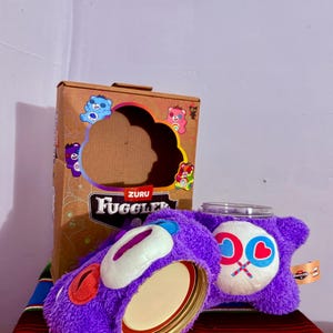 May include: A purple plush Fuggler jar cover with a white face, red and blue accents, and a glass jar. The box behind it has colorful bear illustrations and the word "Fuggler".