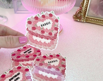 Könnte beinhalten: Drei herzförmige Kuchenaufkleber in Rosa und Weiß mit dem Text "Birthday Girl" und "Baddie" darauf. Die Aufkleber befinden sich auf einer glitzernden rosa Oberfläche.