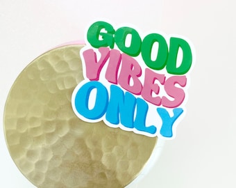 May include: A white sticker with the text "GOOD VIBES ONLY" in colorful, stylized letters. The sticker is on a gold, textured surface.