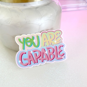 May include: A colorful sticker with the text "You Are Capable" in a rainbow-like font with white outlines. The sticker has a glossy finish and is on a white glittery surface.