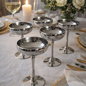 Peut inclure: Mise en place avec cinq coupes à champagne argentées. Une bougie allumée, un verre de vin blanc et un bouquet de roses blanches sont également sur la table. La table est dressée avec des couverts et des assiettes dorés.