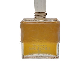 May include: A clear glass bottle of Molinard De Molinard Eau De Parfum. The bottle has a textured surface and a white cap with a brown string.