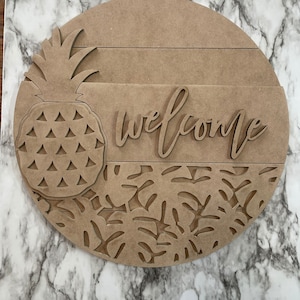 Può includere: Un cartello rotondo in legno con un design ad ananas e la scritta "welcome" intagliata. Il cartello presenta un ananas sul lato sinistro e foglie tropicali in basso. Il cartello è in legno marrone chiaro.