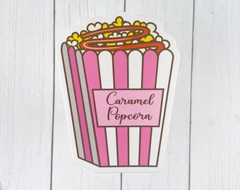 May include: A sticker featuring a cartoon illustration of a popcorn container filled with popcorn and caramel drizzle. The container is pink and white striped with a label that reads "Caramel Popcorn".