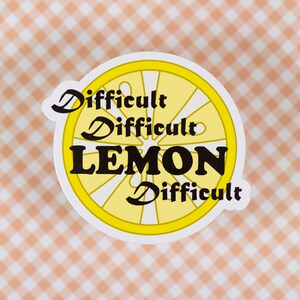 Könnte beinhalten: Ein runder Aufkleber mit einem Zitronenscheiben-Design und den Worten "Difficult Difficult LEMON Difficult" in Schwarz. Die Zitronenscheibe ist gelb mit weißen Details. Auf einem pfirsichfarbenen und weißen Vichy-Hintergrund.