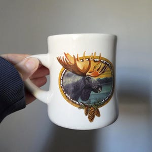May include: White ceramic mug featuring a detailed illustration of a moose with large antlers, set within a circular frame. The moose is depicted against a backdrop of a lake and forest. The mug has a curved handle and a pine cone design.