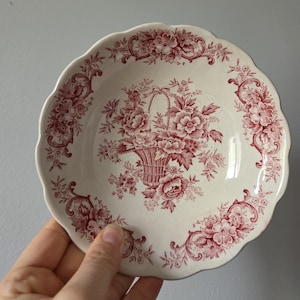 Może przedstawiać: Mały, kremowy ceramiczny talerz z falistą krawędzią. Talerz ma czerwony wzór kosza kwiatowego pośrodku, otoczony kwiatowymi i kręconymi wzorami. Talerz jest trzymany przez dłoń.