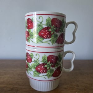 Può includere: Due tazze in ceramica bianca impilate, ciascuna ornata da un motivo a fragole. Il design presenta fragole rosse, foglie verdi e un sottile bordo rosso. Le tazze hanno manici curvi unici.