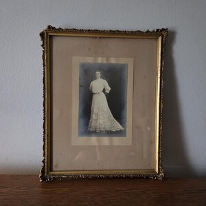Peut inclure: Un cadre rectangulaire doré et orné contenant une photographie vintage en noir et blanc d'une personne debout dans une longue robe blanche à manches bouffantes et jupe à étages, sur fond sombre avec un passe-partout de couleur crème.