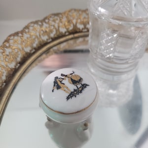 May include: A small white porcelain trinket box with a gold trim and a black and gold design of a couple dancing. The box has three gold feet.