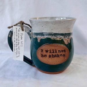 May include: A green ceramic mug with a white rim and a brown oval sticker that says "I will not be shaken". The mug has a handle and a quote from Psalm 62:1 on a white tag attached to the handle.