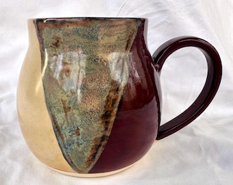 Puede incluir: Una taza grande, redonda y de cerámica con asa. La taza está decorada con un esmalte marrón, verde y crema. La taza es una pieza única de cerámica hecha a mano.