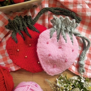 Peut inclure: Deux pochettes tricotées en forme de fraise, une rouge et une rose, avec des tiges et des cordons verts. La pochette rouge a des accents de perles dorées. Un bol de fraises fraîches, de la laine rouge et rose et un petit bouquet de fleurs sont également présents.