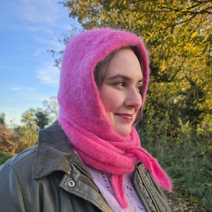 Könnte beinhalten: Eine Frau trägt eine leuchtend pinkfarbene Kapuze und einen Schal. Die Kapuze und der Schal sind aus einem flauschigen Material gefertigt. Sie trägt außerdem einen hellvioletten Strickpullover und eine grüne Jacke.