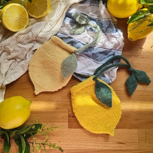 May include: Two knitted lemon-shaped tea bag holders, one yellow and one light yellow, each with green leaf accents and a green braided handle. Fresh lemons and lemon slices are arranged around the holders on a wooden surface, with sprigs of greenery and a white and blue striped cloth.