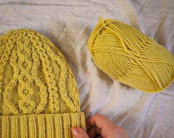 May include: A yellow knitted beanie hat with a cable knit pattern and a matching ball of yarn.