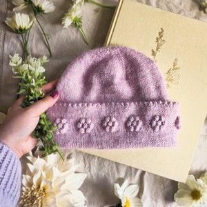 以下が含まれることがあります： 質感のあるデザインの柔らかなパープルのニット帽。帽子のつばは広く、小さな隆起した円形で飾られています。