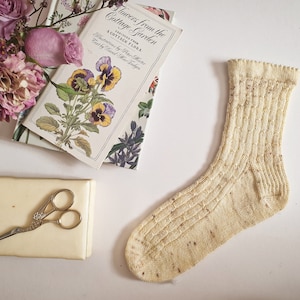 May include: A pair of cream-coloured knitted socks with a textured pattern. The socks are lying on a white surface next to a book and a small box with scissors.