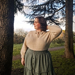 Puede incluir: Una mujer con un suéter de punto color crema con un lazo y una falda verde de capas con lunares blancos. Está de pie en un bosque con árboles en el fondo.