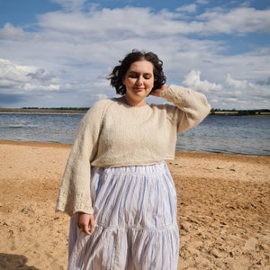 May include: A woman wearing a cream-colored knitted sweater with a white and blue striped skirt. She is standing on a sandy beach with a body of water in the background.