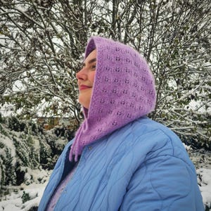 Könnte beinhalten: Eine Person trägt einen lilafarbenen gestrickten Kapuzen-Schal mit einer blauen gesteppten Jacke in einer verschneiten Umgebung.