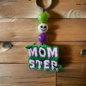 May include: Green, white, and purple bead keychain with a green and white "MOMSTER" sign with black bats.