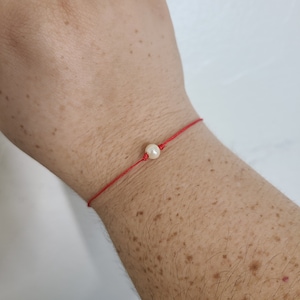 Puede incluir: Un brazalete de cuerda roja con un solo dije de perla blanca.