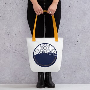 May include: White tote bag with a navy blue mountain and sun graphic. The bag has yellow handles and the text "Some people cross your path and change your whole direction."