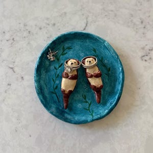 Op de afbeelding: Een ronde, blauwgroene keramieken schaal met twee handbeschilderde, driedimensionale otters die op hun rug liggen. Elke otter houdt een zilverkleurige ring om zijn lichaam. Een paar kleine zilveren oorknopjes ligt op de schaal.