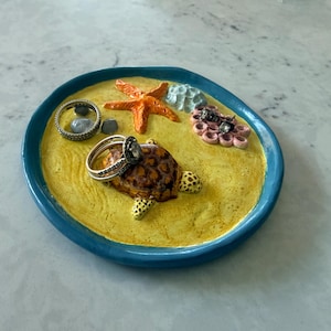 May include: A round ceramic trinket dish with a teal rim and a textured yellow base resembling sand. The dish features a sculpted brown turtle, an orange starfish, a light blue coral piece, and a pink cluster of shells, with two silver rings.