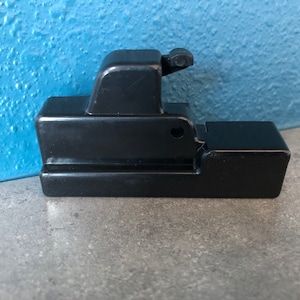 May include: Black plastic latch with a small lever. The latch has a rectangular shape with a rounded edge.