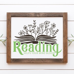 Puede incluir: Cartel enmarcado en madera con fondo blanco. El cartel presenta una ilustraci&oacute;n en negro de un libro abierto con flores y las palabras "Reading Helps Your Mind Bloom" en verde.