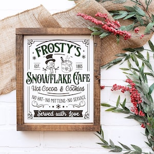 May include: A framed wooden sign with the text "Frosty's Snowflake Cafe" in green and black. The sign includes illustrations of a snowman and a mug of hot cocoa. The sign reads "Hot Cocoa & Cookies" and "Served with Love."