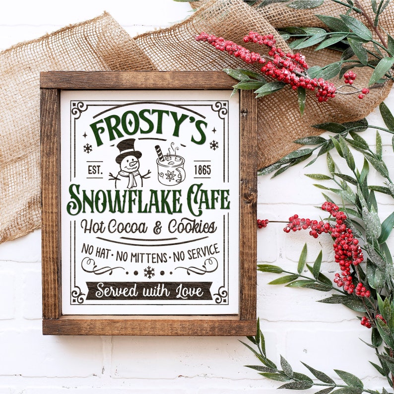 May include: A framed wooden sign with the text "Frosty's Snowflake Cafe" in green and black. The sign includes illustrations of a snowman and a mug of hot cocoa. The sign reads "Hot Cocoa & Cookies" and "Served with Love."