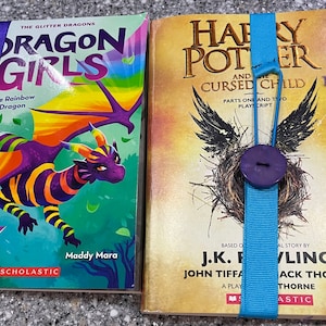Peut inclure: Deux livres broch&eacute;s avec des couvertures color&eacute;es et des signets en ruban bleu. Le livre de gauche est intitul&eacute; "Dragon Girls" avec une illustration de dragon arc-en-ciel. Le livre de droite est intitul&eacute; "Harry Potter et l'Enfant Maudit" avec une illustration sombre d'un nid d'oiseau.